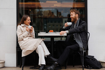 Happy couple drinking coffee and laughing talking to each other sitting at the table of outdoor cafe on city streets