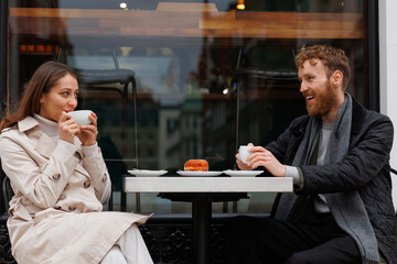 Happy couple drinking coffee and laughing talking to each other sitting at the table of outdoor cafe on city streets