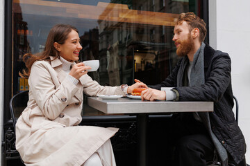Happy couple drinking coffee and laughing talking to each other sitting at the table of outdoor cafe on city streets