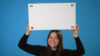 Smiling young business woman in black shirt posing isolated on solid blue background holding white blank sign board with tracking points for text logos.