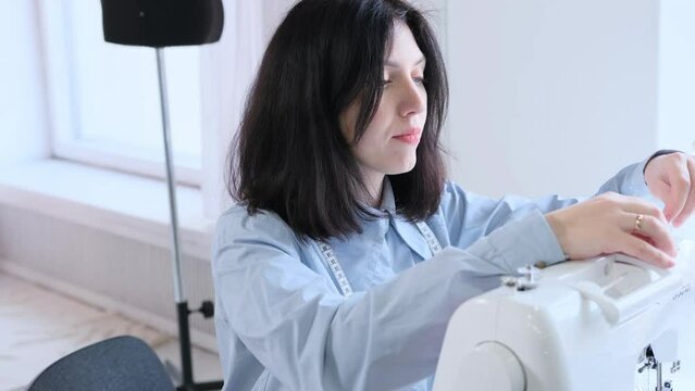 a young Caucasian woman seamstress in a blue shirt and a centimeter on her neck tucks threads into a sewing machine.