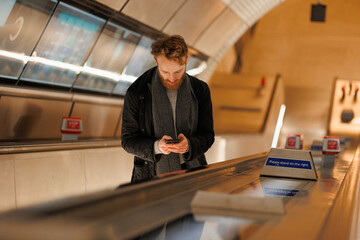Fototapeta premium Bearded man using smartphone while standing on an escalator in the subway