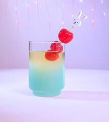 A Pastel Spring Easter Cocktail: Dreamy Green & Yellow Drink Beverage with Three Maraschino Cherries, an Easter Bunny Rabbit Swizzle Stick with Festive Lights on a Light Purple Background