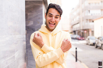 Obraz premium young hispanic man smiling cheerfully and celebrating, with fists clenched and arms crossed, feeling happy and positive