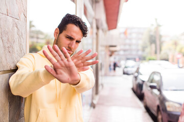 young hispanic man covering face with hand and putting other hand up front to stop camera, refusing...