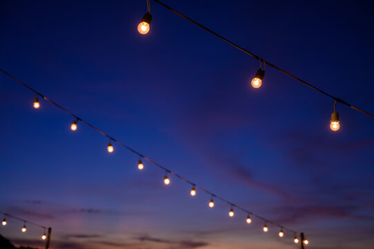 Festoon String Lights Decoration At The Party Event Festival Against Sunset Sky. Light Bulbs On String Wire With Copy Space. Outdoor Holiday Background.