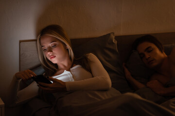 Obraz premium unfaithful woman using smartphone next to sleeping boyfriend at night, cheating concept.