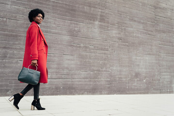 Stylish afro woman holding handbag and wearing a red coat while walking outdoors on the street.