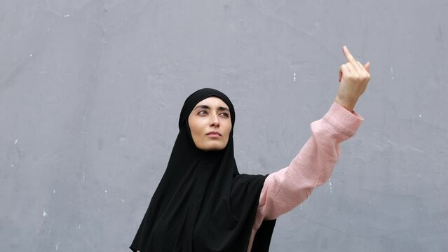 Brash, brave Muslim woman in hijab with hand stretched high upwards provocatively shows her middle finger in protest. Arab Islamic headscarf-covered woman shows middle finger, gesture of middle finger