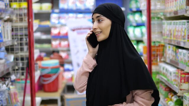 Young Muslim woman in a hijab in the middle of the store shelves affably answers an incoming call. She holds a mobile phone in her hands and smiles when talking with an interlocutor.