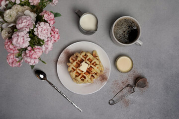 Top view of tasty Waffles Plate, Caramel Sauce, Coffee Cup, Milk, dessertspoon, strainer, pink flowers on a Grey concrete Background