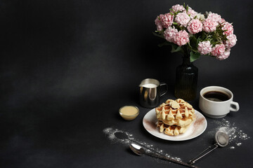 Tasty Waffles Plate, Caramel Sauce, Coffee Cup, Milk, dessertspoon, strainer, pink flowers on a Black concrete Background