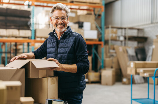 Mature man smiling at the camera while packing cardboard boxes in a warehouse