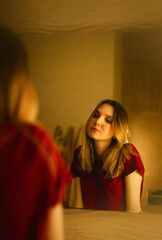 Portrait of a beautiful young blonde woman in a red T-shirt, who is at home in the evening in the bathroom standing at the mirror and admiring herself. Acceptance of your appearance. Self-love.