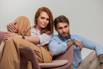 Fashionable beautiful couple in stylish clothes are sitting in a chair on a white background. Beautiful redhead young girl and handsome brutal hipster guy
