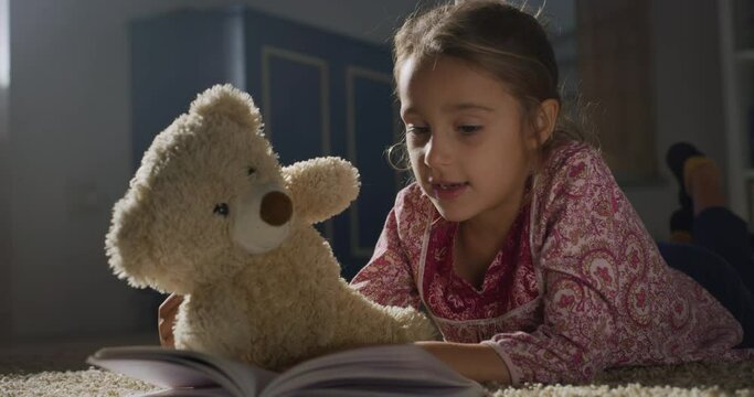 Portrait of a Little Female Child Reading a Book to her Favorite Teddy Bear During Evening. Happy Cute Girl Playing with her Toy, Telling a Story. Concept of Childhood with Cosy Atmosphere
