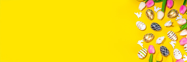 Bright Easter greeting card background. Trendy colored black, golden, stripe painted Easter eggs with white bunny rabbits decor, tulip flower on high-colored yellow background, flat lay copy space