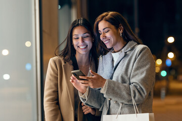 Two beautiful friends talking and using smartphone while walking on the city street with shopping bag.