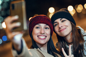Two joyful woman taking a selfie with smartphone while showing peace gesture in the city at night