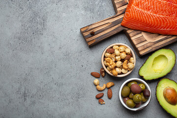 Food sources of healthy unsaturated fat and omega 3: fresh raw salmon fillet, avocado, olives, nuts on cutting board, rustic stone background top view. Healthy nutrition and keto diet, space for text