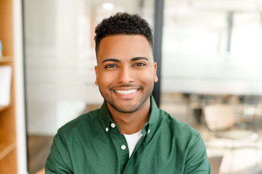 Portrait of happy satisfied Indian young businessman standing in the office and looking at the camera with light smile. Profile photo of cheerfal male office employee, small buissnes owner