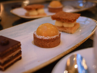 Close up of traditional French mini lemon tart with pie crust in a plate with another mini dessert in background. It's a crisp buttery crust with a smooth tangy lemon custard dessert