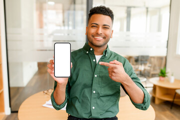 Happy smiling Indian businessman holding smartphone in hand and points finger at empty phone...