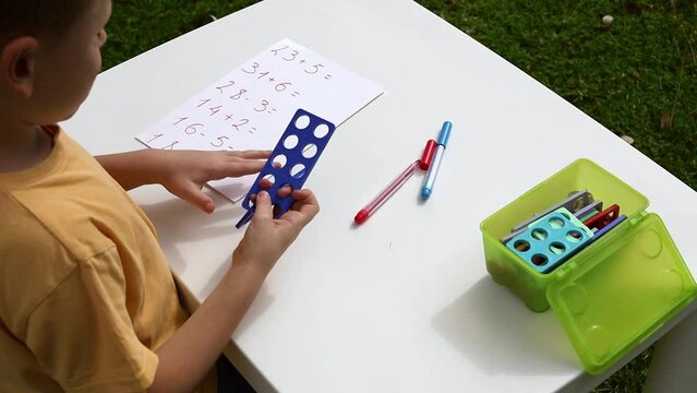 Child with autism has math therapy lesson: uses colorful Numicon shapes, counts, writes answers on paper; sits at white table in sunny garden with bright green grass
