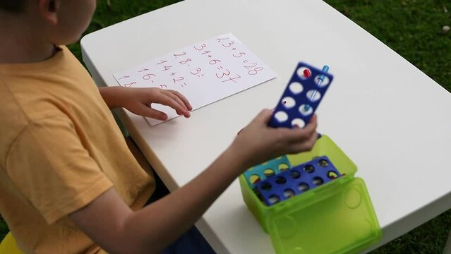 Child with autism has math therapy lesson: uses colorful Numicon shapes, counts, writes answers on paper; sits at white table in sunny garden with bright green grass