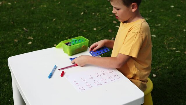 Child with autism has math therapy lesson: uses colorful Numicon shapes, counts, writes answers on paper; sits at white table in sunny garden with bright green grass