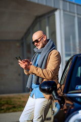 Mature man wearing casual clothes and text messaging while standing outside beside his car
