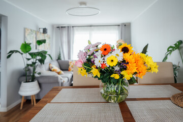 Fototapeta na wymiar Kitchen counter table with focus on vase with huge multicolor various flower bouquet with blurred background of modern cozy living room with couch and green plants. Open space home interior design.