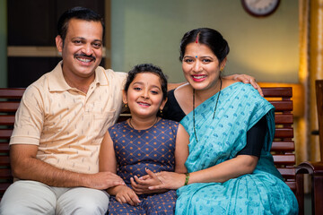 Happy smiling couple with daughter looking camera while sitting on sofa at home - concept of...
