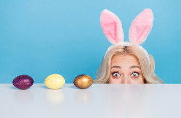 Photo of cute funky woman wear bunny headband hiding table looking eggs isolated blue color...