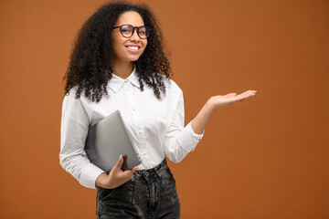 Obraz na płótnie Canvas Beautiful African american business woman carrying laptop and pointing with hand aside, holding empty space above hand palm standing isolated on brown, female office employee presenting, advertising