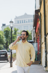 Happy brunette man in sunglasses talking on smartphone while walking on street in Kyiv.