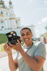 carefree african american tourist with vintage camera smiling near blurred St Andrews Church in Kyiv.