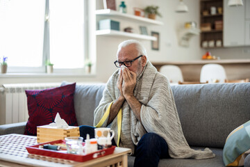 Senior man suffering from flu while sitting wrapped in a blanket on the sofa at home. Elderly Man blowing his nose while lying sick in bed at home. Man with a cold lying in sofa holding tissues