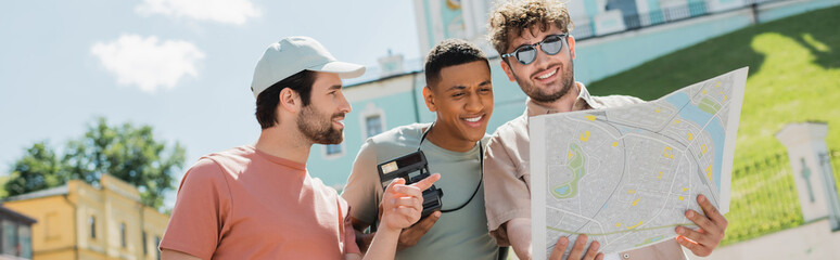 tourist in sun cap pointing at city map near smiling tour guide and african american man on Andrews...