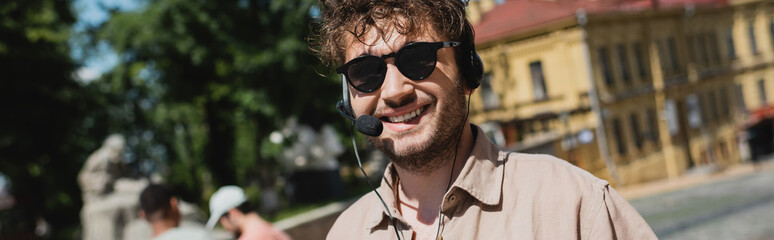 portrait of carefree tour guide in sunglasses and headset smiling at camera on blurred Andrews...