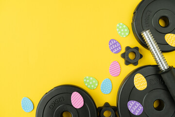 Dumbbells barbell weight plates and Easter eggs decorations. Healthy fitness lifestyle composition. Gym workout and sport training flat lay concept with copy space on yellow background.