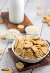 Small round crackers with oatmeal and sesame seeds in a white ceramic bowl on a wooden board on the table. Crispy snack made from wheat flour. in the background is a bottle of milk. Close-up