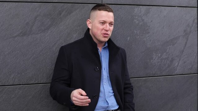 Handsome man in black coat confidently talk standing near building wall. Business man emotionally tell something.