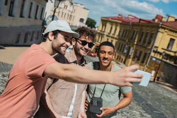 happy tourist in sun cap taking selfie with multiethnic men during excursion on Andrews descent in Kyiv.