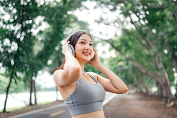 Close up happy asian woman adjusting wireless headphones before starting jogging and listening to music on running route surrounded by nature