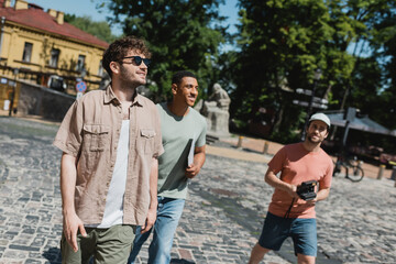 Fototapeta premium smiling multiethnic tourists with vintage camera and map walking with tour guide on Andrews descent in Kyiv.