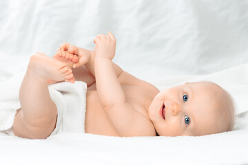 Cute happy baby with blue eyes lying on white bed and holding legs. Little smiling boy looking at camera. Child playing with his feet.