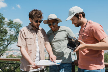 young tour guide in sunglasses looking at travel map near interracial tourists with vintage camera in city park.