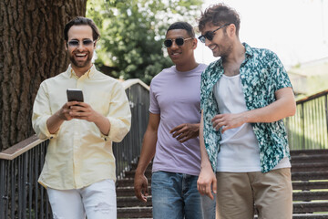 Cheerful man using smartphone while walking near multiethnic friends on stairs in summer park.