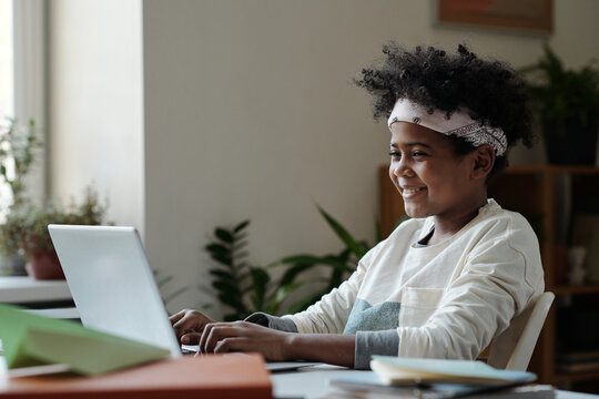 Cute smiling schoolchild sitting by desk in front of laptop and typing while carrying out home assignment or communicating with friends